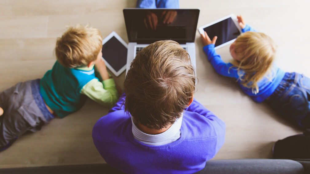 Zwei Kinder und der Vater vor einem Laptop und Tablets
